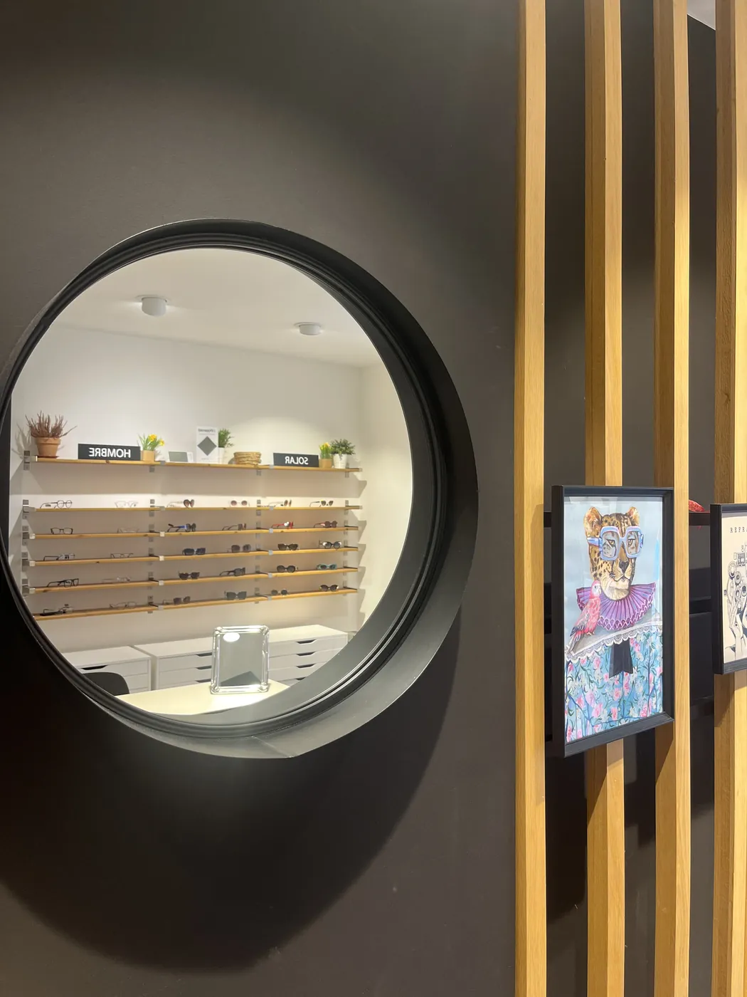 Espejo redondo de marco negro en el interior de la óptica, con el reflejo de los expositores de monturas y de la celosía de listones de madera