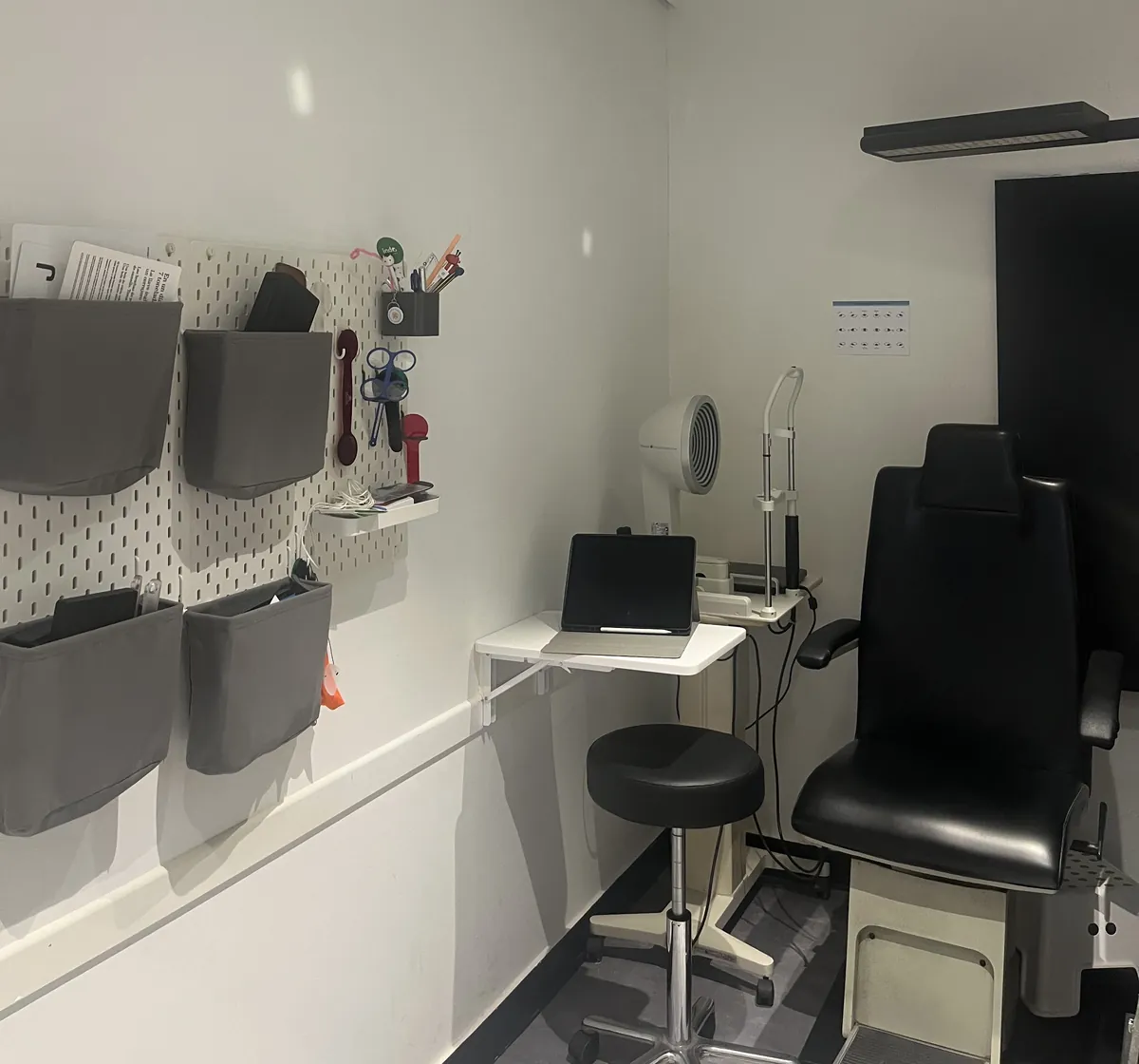 Gabinete de exámenes visuales: sillón, taburete y mesa con una tablet, junto a un organizador de pared