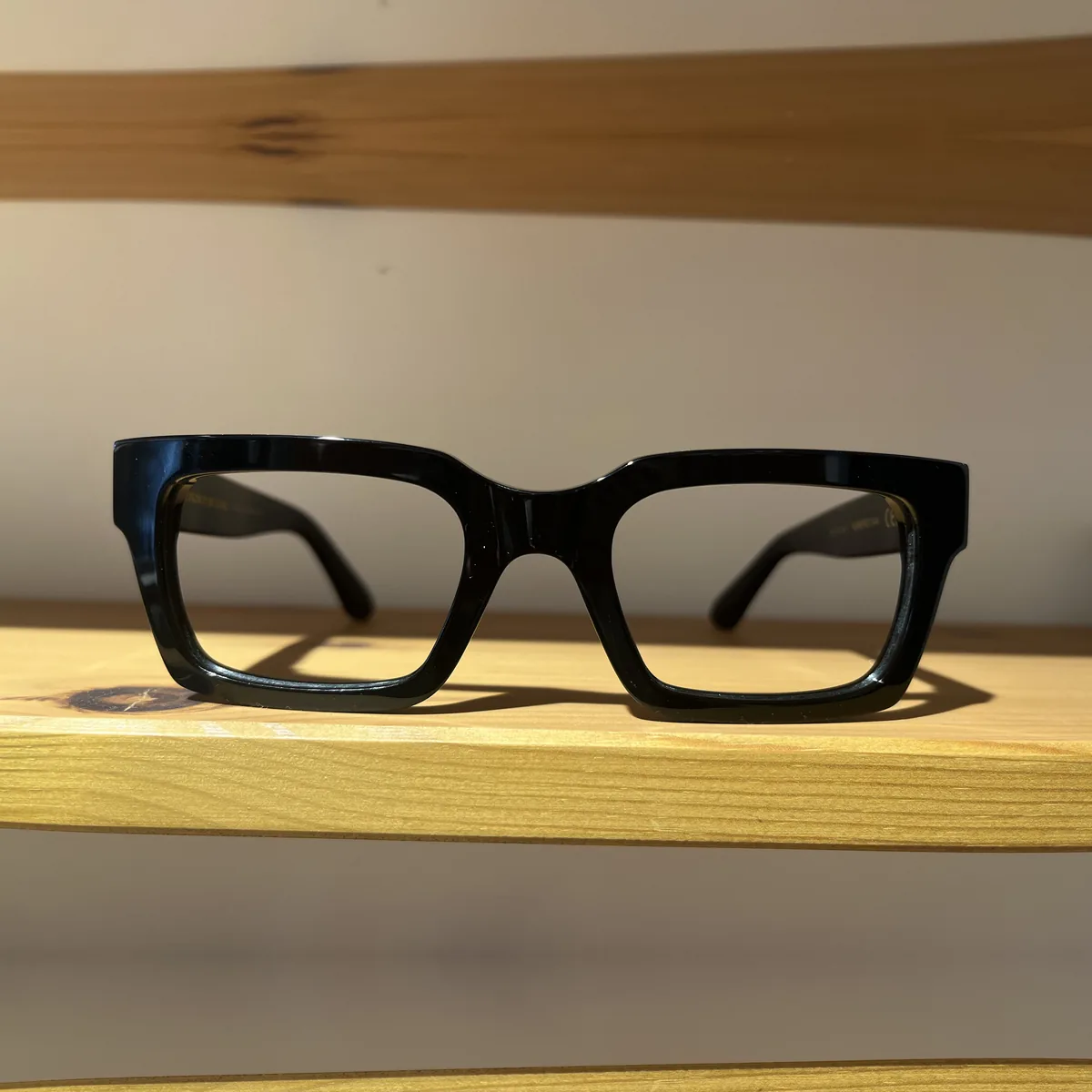 Montura de pasta negra de forma rectangular y línea gruesa, sobre una balda de madera clara