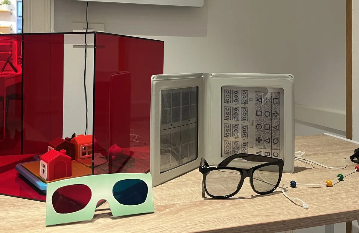 Material de terapia visual sobre la mesa: una caja roja translúcida con casitas dentro, unas gafas de filtro rojo y azul, láminas de test y una montura de prueba
