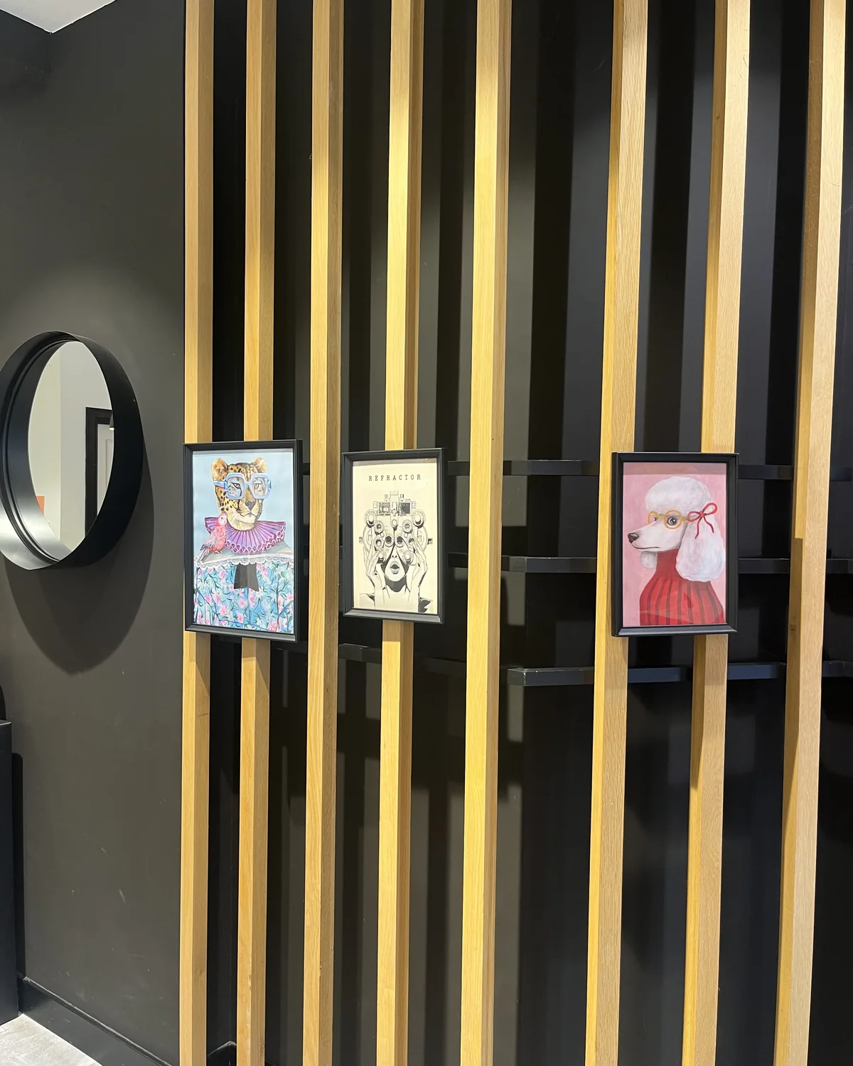 Pared de listones de madera clara sobre fondo negro, con un espejo redondo y tres láminas enmarcadas